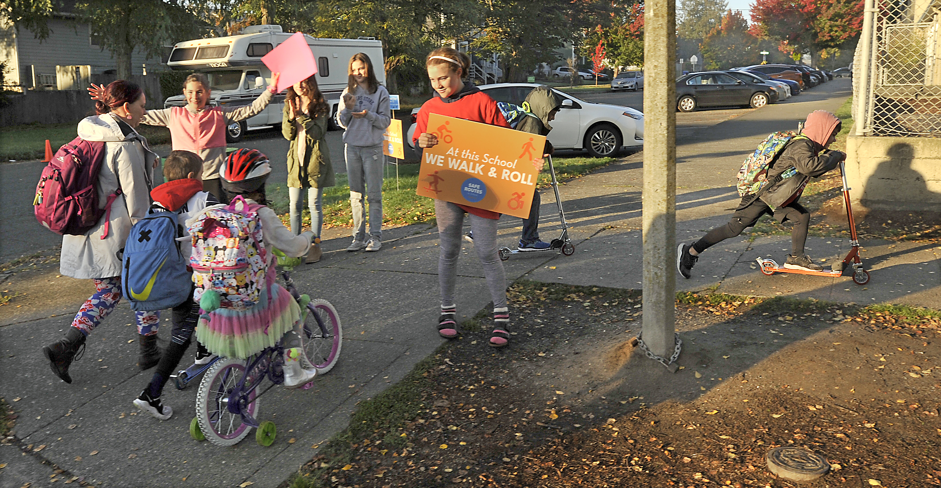 students walking and scooting to school with an adult celebrating them, holding a sign saying At this School we Walk and Roll